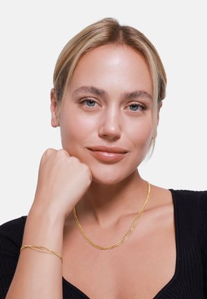 Gouden vergulde gedraaide ketting- en armband set met een stijlvolle, gestructureerde oppervlak, gedragen door een model met een gladde huid en lichte make-up, tegen een witte achtergrond.