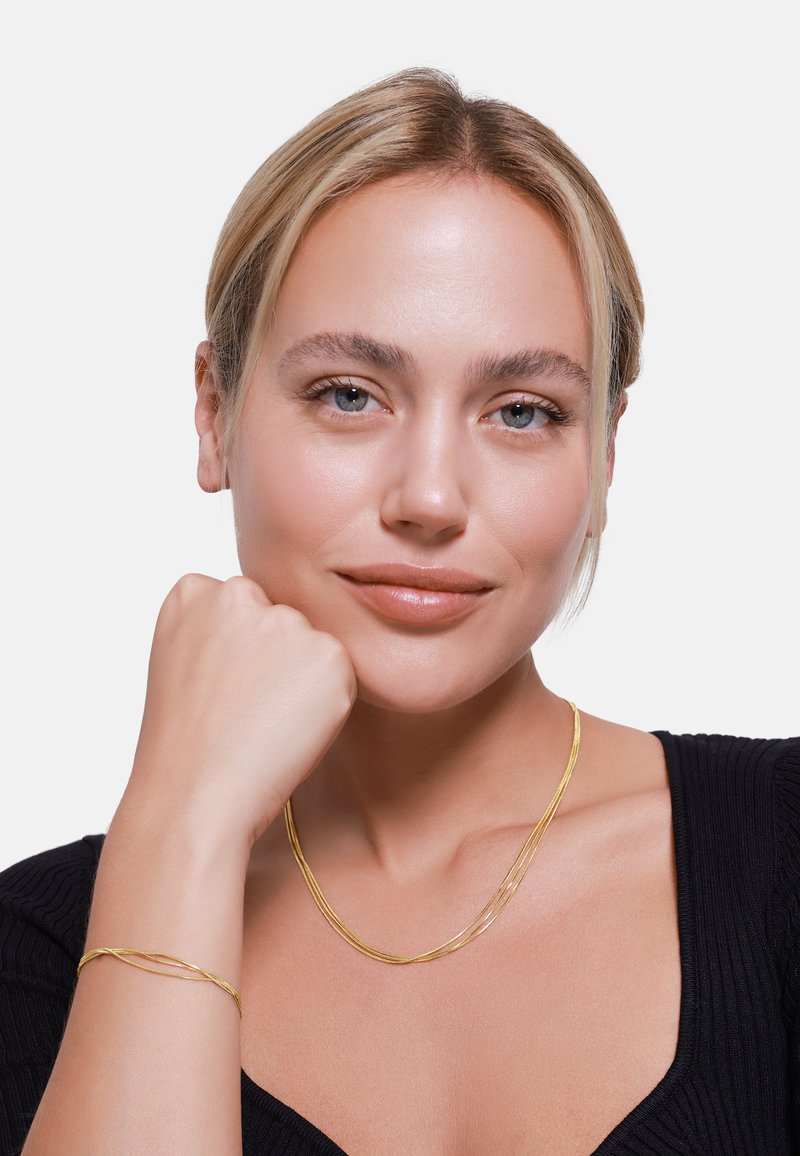 Set di collana e bracciale intrecciati in tono oro con superficie liscia e texture, indossato da un modello con pelle liscia e trucco leggero, su sfondo bianco.