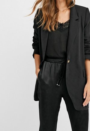 Blazer noir avec un seul bouton doré, accompagné d'un haut noir avec des dentelles et d'un pantalon noir à cordon avec une texture lisse.