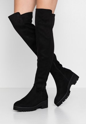 Bottes noires en daim au-dessus du genou avec des talons bas et épais portées sur des jambes croisées sur un fond clair uni.
