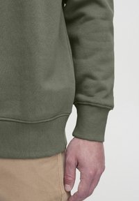 Olivzaļš džemperis ar mīkstu tekstūru, ribota aproce un taisna apakšmala, kopā ar haki biksēm, rokas atpūšas sānos.