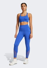 Modrý športový podprsenka a legíny s vysokým pásom s sieťovinovými detailmi. Hladká, elastická látka s priliehavým strihom. Biele tenisky sú súčasťou outfitu.