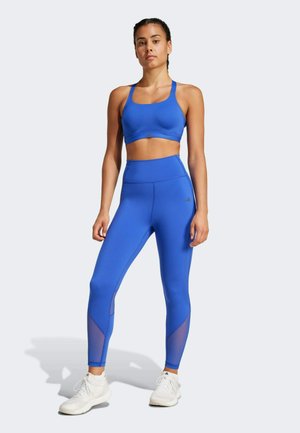 Modrý športový podprsenka a legíny s vysokým pásom s sieťovinovými detailmi. Hladká, elastická látka s priliehavým strihom. Biele tenisky sú súčasťou outfitu.