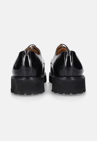 Een paar glanzende zwarte leren dresschoenen met dikke, robuuste zwarte rubberen zolen, gezien van achteren op een witte achtergrond.