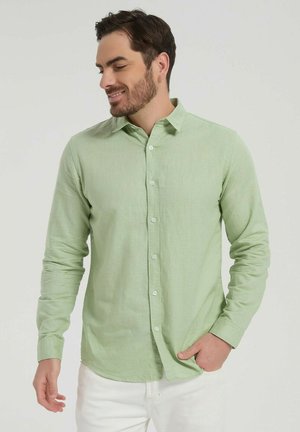 Homme barbu portant une chemise à manches longues vert clair boutonnée et un pantalon blanc, souriant et regardant vers sa gauche.