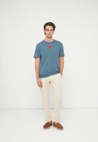 Camiseta azul de algodón con mangas cortas y un parche de logo rectangular rojo, combinada con pantalones de color crema y zapatos marrones sin cordones.