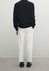 Homme de dos portant un pull noir, un pantalon blanc et des chaussures noires, debout sur un sol et un mur d'arrière-plan gris neutre.