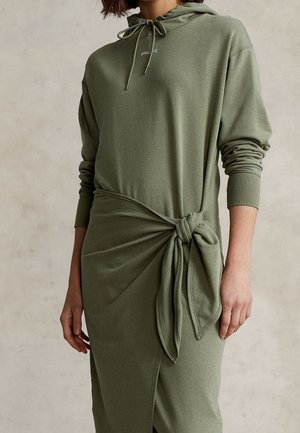 Mujer vistiendo un vestido con capucha de color verde oliva, de mangas largas, con un nudo frontal y un sutil logo RLX en el pecho.