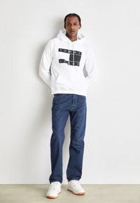 Sudadera blanca con el logo negro "TOMMY JEANS", combinada con jeans de mezclilla oscura y zapatillas blancas con suelas naranjas. Textura de tela suave.