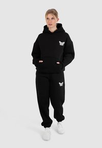 Sort hættetrøje med frontlomme og hvidt sommerfuglelogo på brystet, kombineret med matchende sorte sweatpants med et lille logo.