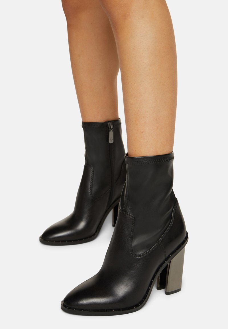 Black ankle boots made of leather with a stretchy upper, pointed toe, and a silver block heel. Features a side zipper and decorative studs.