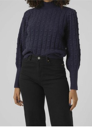 Femme portant un pull en maille torsadée bleu marine foncé à manches longues et un jean noir taille haute sur un fond gris clair uni.