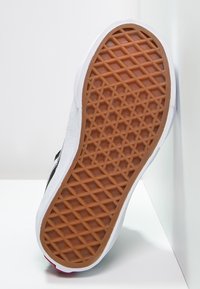 Semelle d'une basket blanche avec une bande de roulement en caoutchouc marron à motif gaufré, appuyée contre un mur blanc d'angle.
