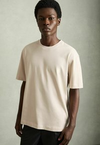 Tricou scurt cu mânecă scurtă, de culoare bej deschis, cu guler rotund, fabricat din bumbac, cu o croială lejeră și textură fină. Purtați cu pantaloni întunecați.