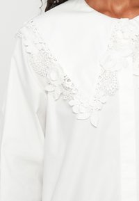 Camicia bianca con ampio colletto triangolare caratterizzato da un intricato ricamo di pizzo floreale lungo i bordi e il scollo.