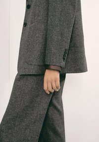 Traje de lana gris con un ajuste entallado, que presenta grandes botones y un acabado texturizado. La mano está posicionada sobre la costura del pantalón.