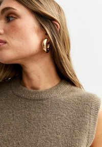 Beige ärmelloses Strickoberteil mit einer strukturierten Optik, kombiniert mit großen glänzenden goldenen Creolen. Das Haar fällt lässig über eine Schulter.