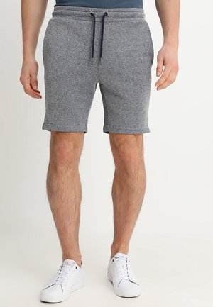 Man draagt grijze shorts met trekkoord en witte sneakers, staand tegen een effen lichte achtergrond.