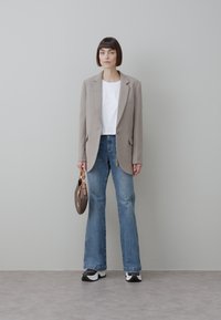 Blazer beige, camisa blanca corta, vaqueros acampanados azul oscuro, zapatillas blancas y negras, con un bolso marrón, sobre un fondo neutro.