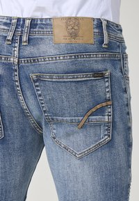 Los jeans de mezclilla azul deslavado presentan un bolsillo trasero en forma de diamante, un parche de cuero color marrón con un diseño de calavera y costuras amarillas.