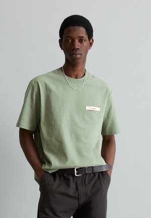Jonge man die een lichtgroen shirt met korte mouwen draagt, met een klein wit logo en zwarte broek met een zwarte riem, handen in de zakken, neutrale achtergrond.