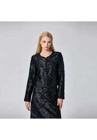 Femme blonde portant une veste longue à manches et motif floral noir ainsi qu'une jupe assortie, devant un fond gris uni.
