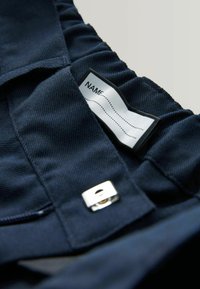 Marineblåt stof med en tekstureret finish, med et påsyet mærke til personalisering og en sølvfarvet hardware-detalje.