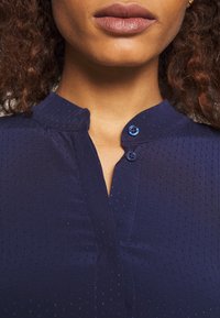 Blouse bleu marine à surface texturée, avec un col rabattable et deux boutons bleus, présentant un design simple et moderne.