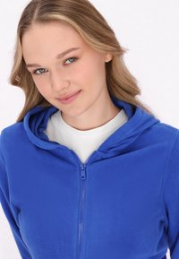 Felpa in pile blu con zip frontale, dotata di cappuccio e texture liscia. Indossata sopra una maglietta bianca.