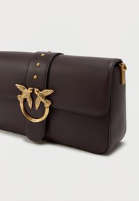 Σκούρα καφέ δερμάτινη τσάντα χειρός με καπάκι κλεισίματος, χρυσό κούμπωμα με διπλά πουλιά και διακοσμητικές μπίλιες στη ζώνη, σε λευκό φόντο.