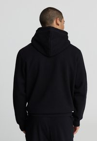 Uomo con capelli corti che indossa un semplice hoodie nero e pantaloni neri, visto da dietro su uno sfondo grigio chiaro.