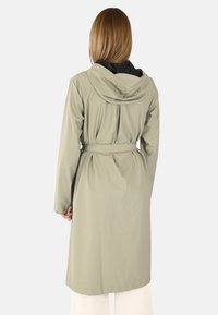 Personne avec de longs cheveux portant un manteau à capuche beige clair ceinturé, vue de dos, se tenant contre un fond uni.