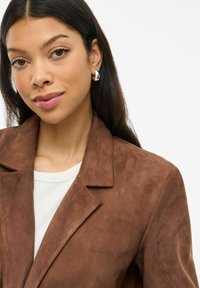 Nuori nainen, jolla on pitkät tummat hiukset, käyttää hopeisia rengaskorviksia, ruskeaa nobukkitakkia ja valkoista paitaa, hymyilee hieman valkoista taustaa vasten.