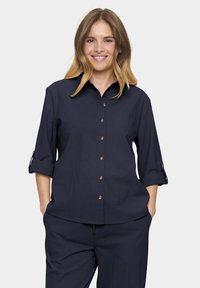 Navy blue button-up shirt with rolled sleeves, featuring brown buttons and a relaxed fit. Soft fabric with a smooth texture.