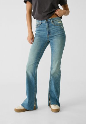 Persoon draagt een high-waisted blauwe jeans met scheuren aan de voorkant van de pijpen en witte sneakers, zwarte T-shirt gedeeltelijk in de broek gestopt.