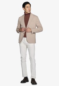 Beige blazer over en rustfarvet turtleneck, parret med hvide bukser og mørke dress sko. Funktioner inkluderer en notch-lapel og tekstureret stof.