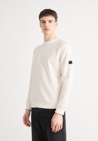 Lichtbeige crewneck sweatshirt van zacht materiaal, met ribbels aan de boorden en onderrand, en een zwart etiket op de linkermouw.