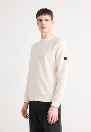 Sweatshirt beige clair à col rond fabriqué en tissu doux, avec des poignets et un ourlet côtelés, et une étiquette noire sur la manche gauche.