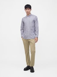 Jeune homme portant une chemise rayée bleu et blanc à boutons, un pantalon beige et des chaussures noires, debout les mains dans les poches.