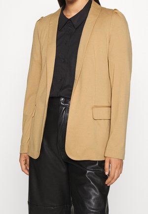 Person trägt einen beigen Blazer über einem schwarzen Hemd mit Knöpfen und schwarzen Lederhosen, steht vor einem schlichten hellen Hintergrund.