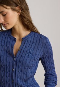 Femme portant un cardigan bleu en maille torsadée avec des boutons dorés et les initiales brodées RL, regardant vers le bas avec une boucle d’oreille dorée visible.
