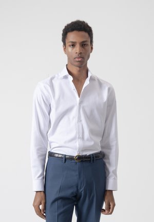 Joven con camisa blanca de vestir metida dentro de pantalones azules con un cinturón negro, de pie contra un fondo liso de color gris claro.