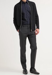 Cardigan nero lavorato a maglia con zip argentata, indossato sopra una camicia blu navy con bottoni. Pantaloni grigi su misura e scarpe da uomo nere completano l'outfit.