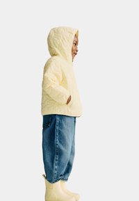 Kind in einer gelben, gesteppten Kapuzenjacke, lockeren blauen Jeans und hellgelben Stiefeln, steht im Profil mit den Händen in den Taschen.