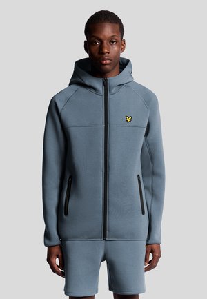 Lichtblauwe hoodie met een ritsfront, twee zwarte ritszakken en een klein geel logo op de borst. Gladde stof met een relaxte pasvorm.