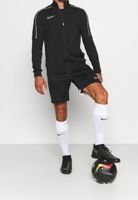Black Nike zip-up jacket with white accents, black shorts, white socks, and black cleats, standing on a black and colorful soccer ball.