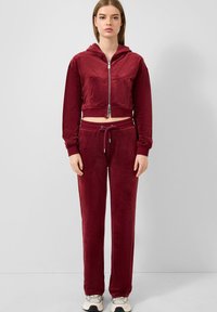 Vrouw in een bordeauxkleurige velours zip-up hoodie en bijpassende jogpants met witte sneakers, staande tegen een effen lichtgrijze achtergrond.
