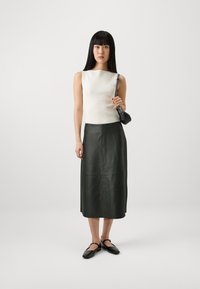 Hvit ribbet ermeløs topp, mørkegrønn skinn midi-skjørt, og svarte Mary Jane-sko. Modell som holder en liten svart veske, stående mot en ensfarget bakgrunn.