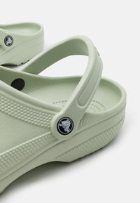 Zoccoli in gomma verde con una parte superiore testurizzata, fori ovali e logo in evidenza. Presentano una fascia flessibile per una vestibilità sicura e comfort.