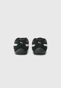 Black Puma sneakers featuring a white stripe and gold logo, viewed from the back, highlighting their sleek design and textured soles.